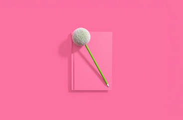 PInk hardcover notebook lying flat on a matching pastel pink background with a green pen on the notebook with a pom pom.  Center composition with copy space and room for text.