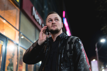 A smiling man wearing in black jacket, is holding scrolling texting in his cellphone. A smiling man calls for a taxi in an app. online, networking, outdoor night city street