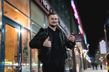 A smiling man wearing in black jacket, is holding scrolling texting in his cellphone. A smiling like. online, networking, outdoor night city street