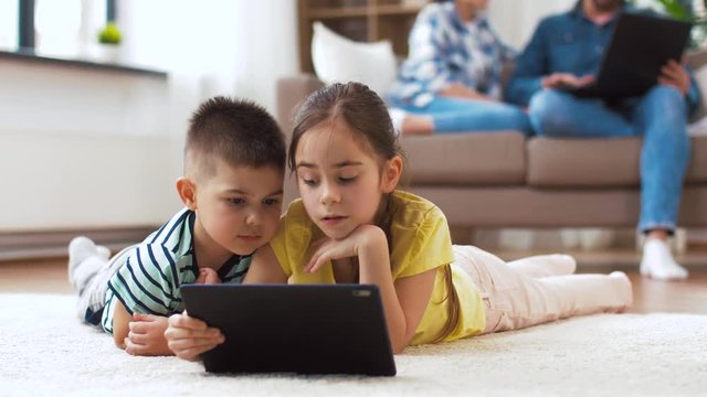 childhood, technology and family concept - little brother and sister with tablet pc computer lying on floor at home
