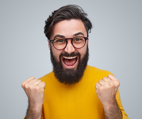 Excited man with beard in glasses standing with fists up wearing yellow sweater on gray background.