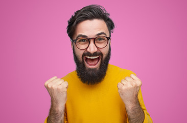 Happy bearded man wearing glasses standing with fists up on pink background. Epic win concept