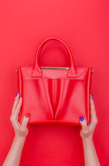 Bright summer fashion ladies accessories. Stylish red leather handbag or shopper bag on a red background. Women hands with bright manicure holding bag from top. Top View. Flat Lay. Copy Space. Mock up