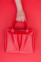 Bright summer fashion ladies accessories. Stylish red leather handbag or shopper bag on a red background. Women hands with bright manicure holding bag from top. Top View. Flat Lay. Copy Space.