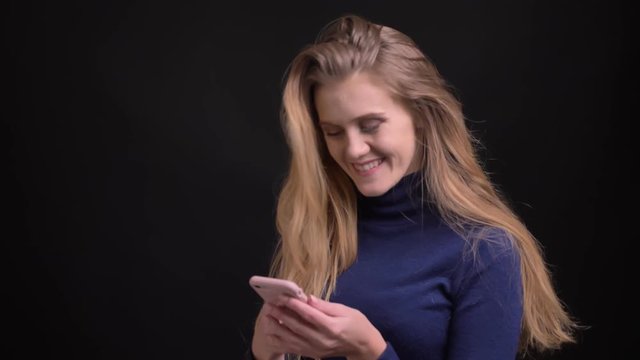 Young blonde model working with smartphone raises glance to camera and smiles into it on black background.