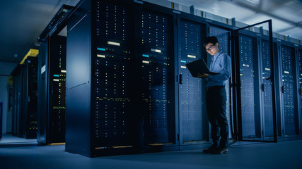 In Data Center: Male IT Technician Running Maintenance Programme on a Laptop, Controls Operational Server Rack Optimal Functioning. Modern High-Tech Telecommunications Operational Super Computer.