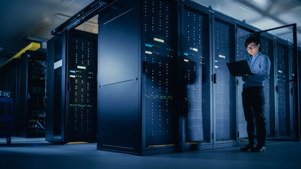 In Data Center: Male IT Technician Running Maintenance Programme on a Laptop, Controls Operational...