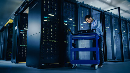 In Data Center: Male IT Technician Running Maintenance Programme on a Laptop, Controls Operational...