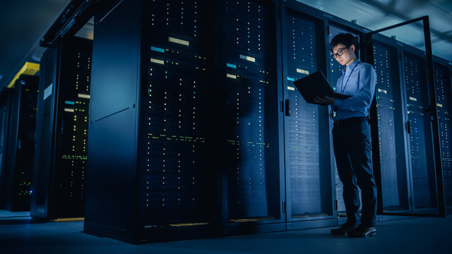 In Data Center: Male IT Technician Running Maintenance Programme on a Laptop, Controls Operational Server Rack Optimal Functioning. Modern High-Tech Telecommunications Operational Super Computer.