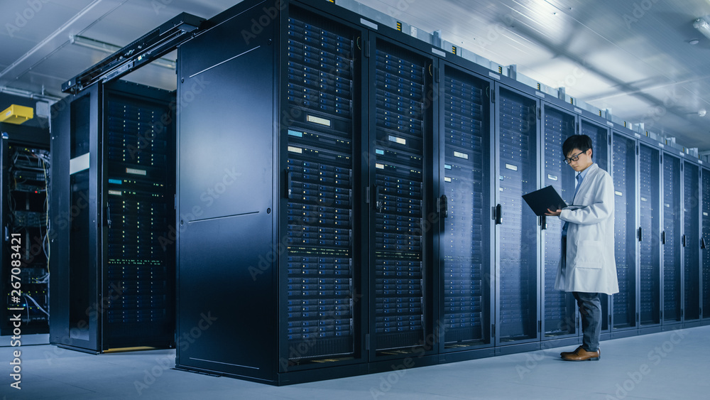 Poster in data center: male it specialist wearing white coat stands beside server rack, uses laptop compute