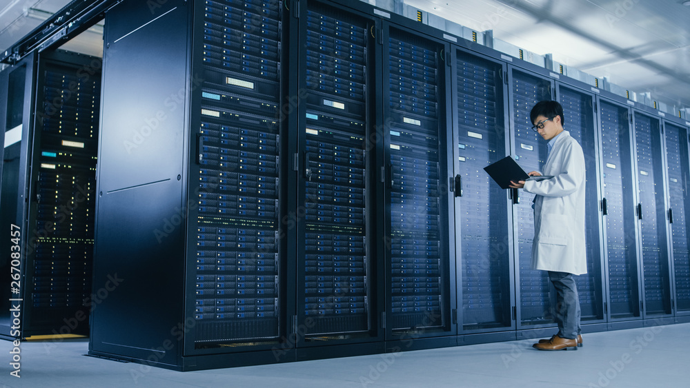 Poster In Data Center: Male IT Specialist Wearing White Coat Stands Beside Server Rack, Uses Laptop Computer to Run Maintenance Diagnostics Tools, Controls so that Database Works at Optimal Level. 