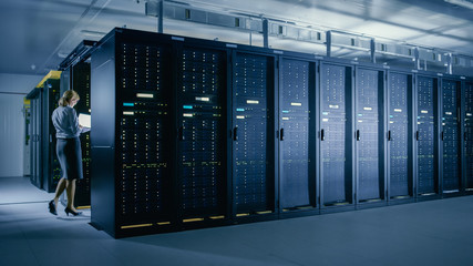 In Data Center: Female IT Technician Walks Between Rows of Server Racks, Uses Laptop Computer to...