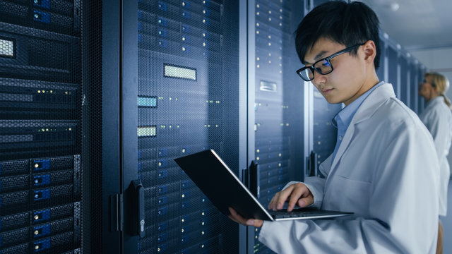 In Data Center: Male and Female IT Specialists Wearing White Coats Work with Server Racks, Use Laptops to Run Maintenance Diagnostics. People wearing Lab Coats Working with Datacenter Database.