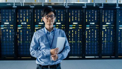 In the Modern Data Center: Portrait of IT Engineer Stands Beside the Row of Server Racks, Finishing...