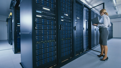 In Data Center: Female IT Technician Stand Near Opened Rack Cabinet and Runs Maintenance Program on Laptop, Controls Operational Server's Optimal Functioning. 