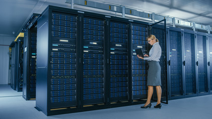 In Data Center: Female IT Technician Stand Near Opened Rack Cabinet and Runs Maintenance Program on Laptop, Controls Operational Server's Optimal Functioning. 
