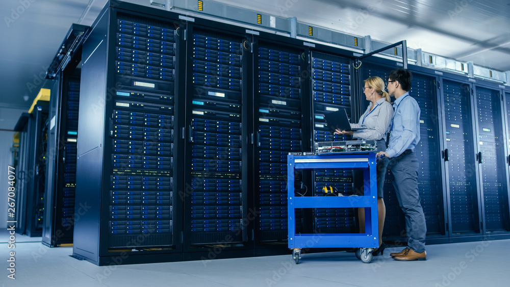 Poster In the Modern Data Center: Engineer and IT Specialist Work with Server Racks, on a Pushcart Equipment for Installing New Hardware. Specialists Doing Maintenance of the Database. Low Angle Shot.
