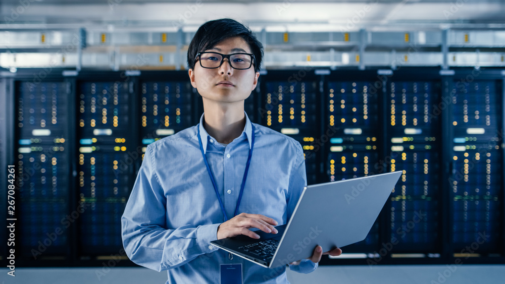 Poster In the Modern Data Center: Portrait of IT Engineer Standing with Server Racks Behind Him, Holding Laptop and Looking at the Camera. Finishing Doing Maintenance and Diagnostics Procedure.