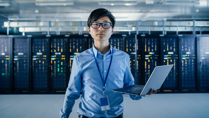 Fototapeta premium In the Modern Data Center: Portrait of IT Engineer Standing with Server Racks Behind Him, Holding Laptop and Looking at the Camera. Finishing Doing Maintenance and Diagnostics Procedure.