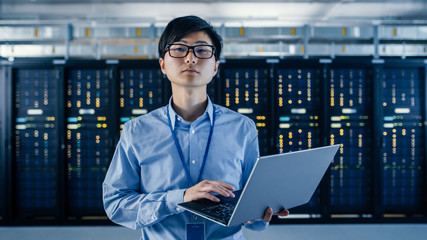 In the Modern Data Center: Portrait of IT Engineer Standing with Server Racks Behind Him, Holding Laptop and Looking at the Camera. Finishing Doing Maintenance and Diagnostics Procedure.