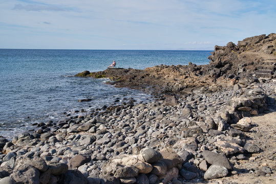 Fisherman sitting on a cape on Playa grande, full of black lava sones in Puerto del Carmen, Lanzarote, Spain