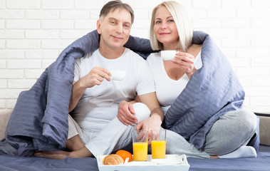 Portrait of happy playful couple relaxing in comfortable cozy bed looking at each other, cheerful man and woman having fun posing. Holding cups of coffee while covering their shoulders with blanket.