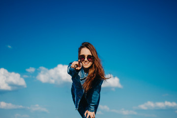 Young woman in sunglasses and denim jacket on blue sky background smiling shows hand to the side