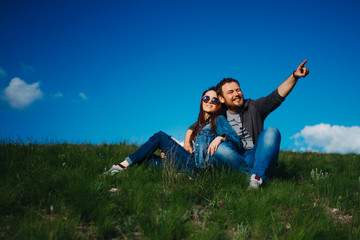 A man and a woman in love are resting in nature, sitting on a green hill against a blue sky hugging. A man shows his hand to the side, woman smiling