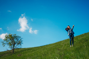 A woman in jeans and a denim jacket stands on top of a hill holding her head from emotion and shows aside