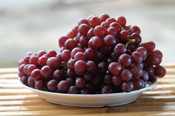 Purple grapes placed in a white plate on a bamboo table, blurred background.