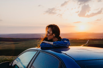 Woman standing leaning on the car at sunset