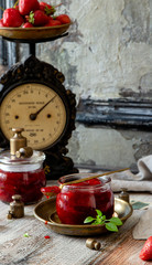 Two glass jars with homemade canned strawberry jam, marmalade, jelly on rustic wooden grey table with basil, lemon, gold weights, bowl of berries, vintage scales, spoons opposite old concrete wall