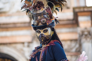 Young girl with long dark braids in a carnival Venetian mask with a closed mouth and a hat with multi-colored feathers, a mechanical Breguet clock and details of a clock mechanism, a chain, precious s