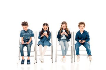 four children in denim clothes sitting on chairs and using smartphones on white