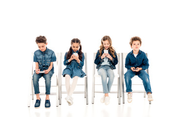 Obraz premium four children in denim clothes sitting on chairs and using smartphones on white