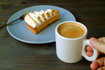 Hand Holding a Cup of Coffee with a Slice of Lemon Meringue Tart in Background 