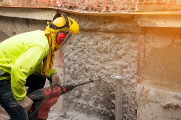 Construction worker Wearing green safety shirt Is using a large drill machine to drill a wall in the area that is building a large building