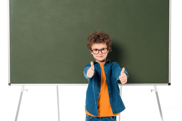 smiling schoolboy in glasses standing near blackboard and showing thumbs up isolated on white