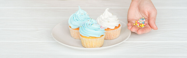 cropped view of woman holding sprinkles in hand and plate with cupcakes on wooden table