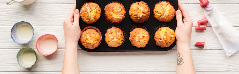 panoramic shot of woman holding baking tin with cupcakes on table with baking tools
