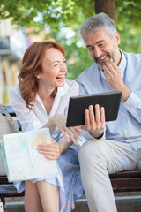 Happy smiling mature tourists sitting on a bench looking at a tablet and laughing. Husband is holding a tablet and wife is holding a city map