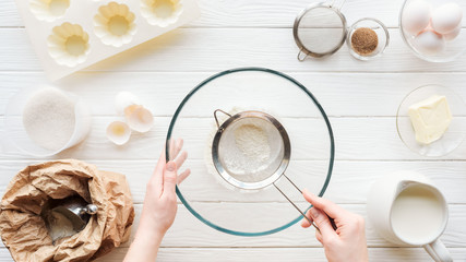 partial view of woman sieving flour in bowl on table with ingredients