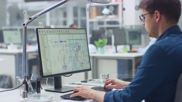 Over the Shoulder Shot of Engineer Working with CAD Software on Desktop Computer, Screen Shows Technical Drafts and Drawings. In the Background Engineering Facility Specialising on Industrial Design