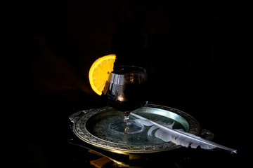 On a silver tray is a glass of red wine with an orange slice on the edge. Nearby is a goose feather. Concept - a mysterious evening