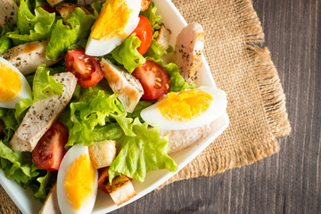 Fresh salad made of tomato, ruccola, chicken breast, eggs, arugula, crackers and spices. Caesar salad in a white, transparent bowl on wooden background