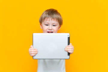 Surprised blonde three years old boy with his mouth open surprised, holding in his hands a tablet pc and looking at the camera on yellow background