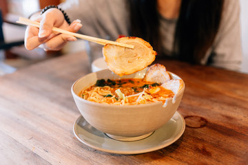 Woman pinching Chashu Pork with chopsticks from Spicy Chashu Miso Ramen: Japanese noodle in vegetable broth mix Miso paste with Chashu pork, sprouts, minced pork, seaweed and half boiled egg.