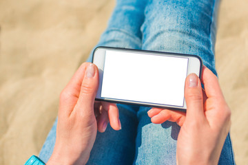 Image Mockup of beautiful hands of a woman holding a white mobile phone with a blank screen on her hip on the beach, on the sand. Close up.