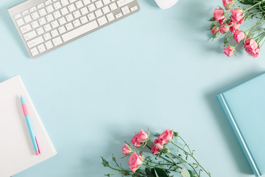 Female home office workplace with keyboard, notepad, rose flowers, notebook on pastel blue background. Business minimal concept for women. Flat lay, top view, copy space