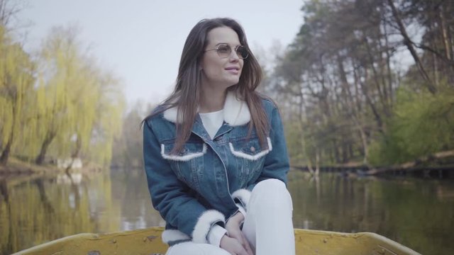 Portrait of a gorgeous young woman in sunglasses and a denim jacket floating on a boat on a lake or river. Beautiful brunette is actively relaxing on a day off or traveling enjoying nature.